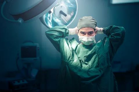 A surgeon in a green suit stands confidently before a bright surgical light, ready for the procedure ahead.
